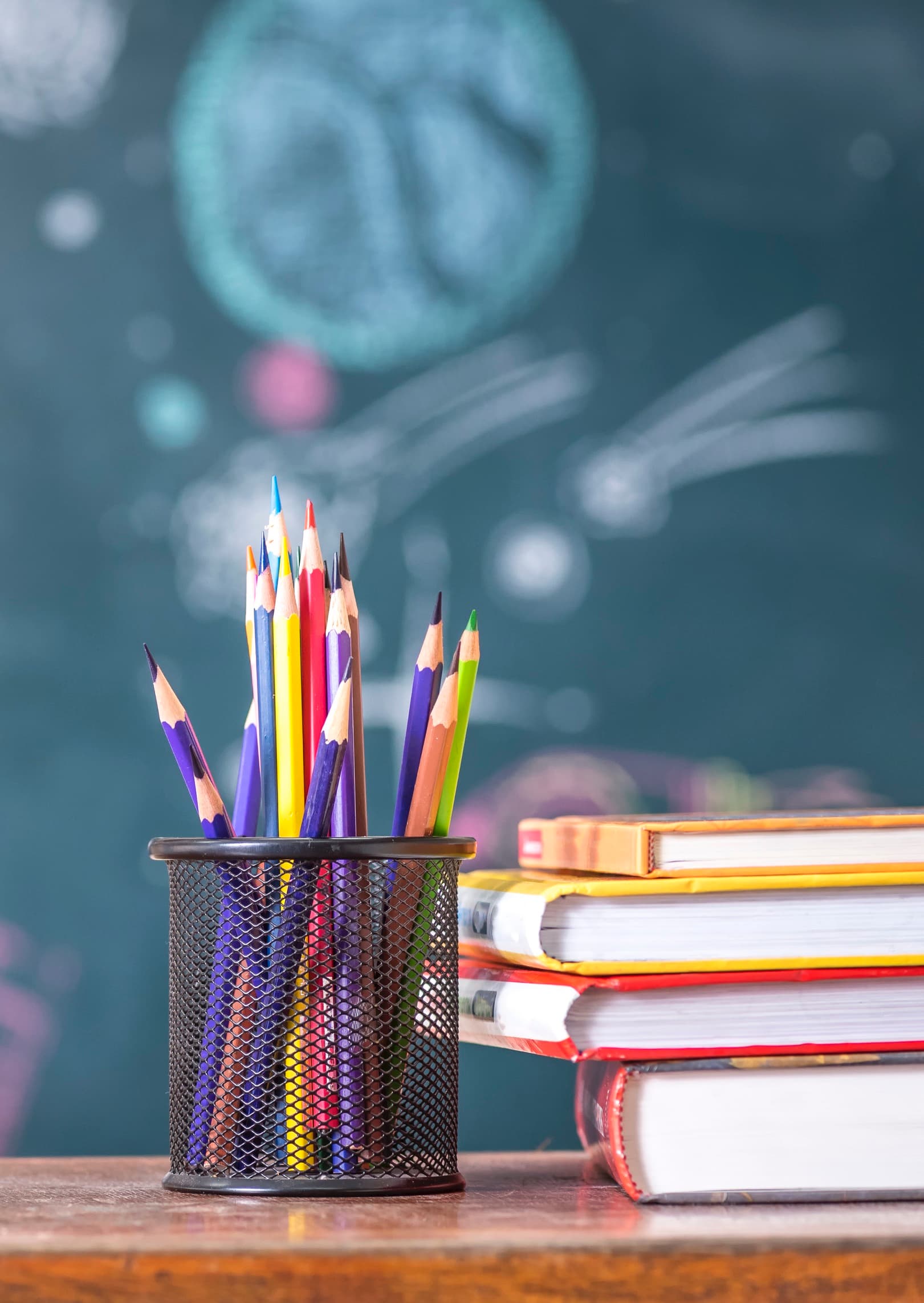 a mesh metal cup of colored pencils sitting next to a stack of school books, with a background of a chalkboard with space images on it