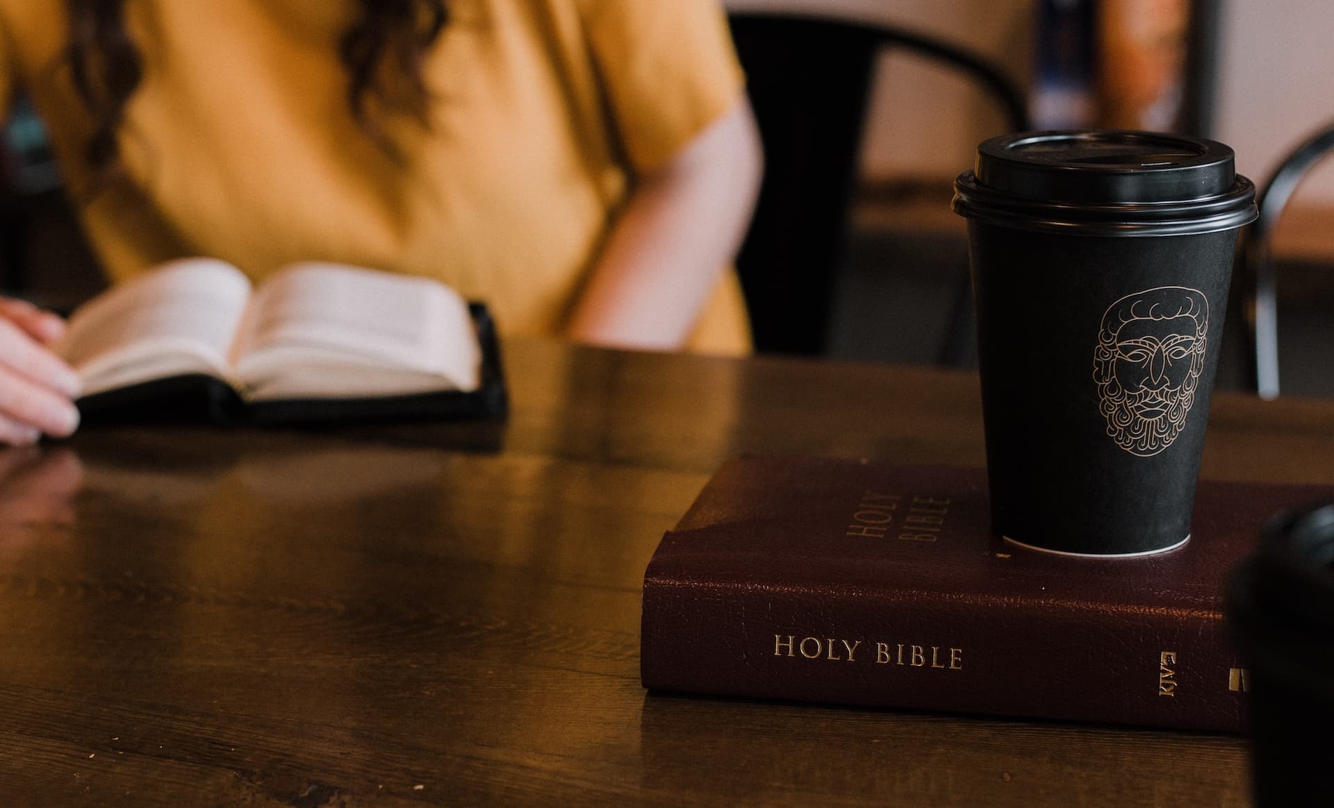 a coffee cup sits on top of a Bible, and another person with an open bible sits in the background