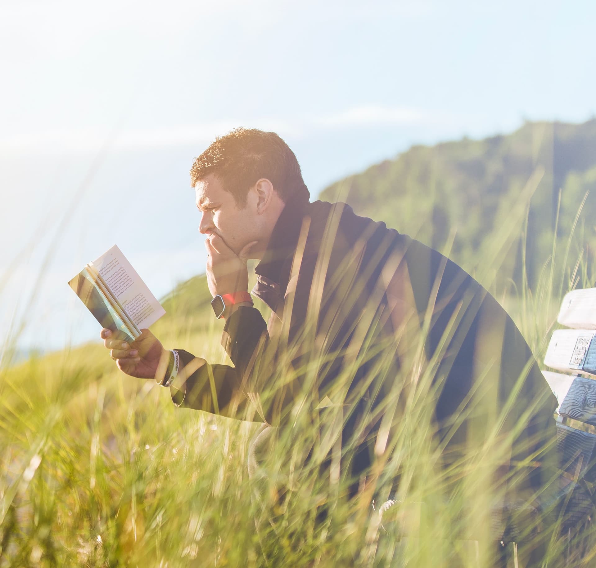 man reading a book on a bench in a grassy field