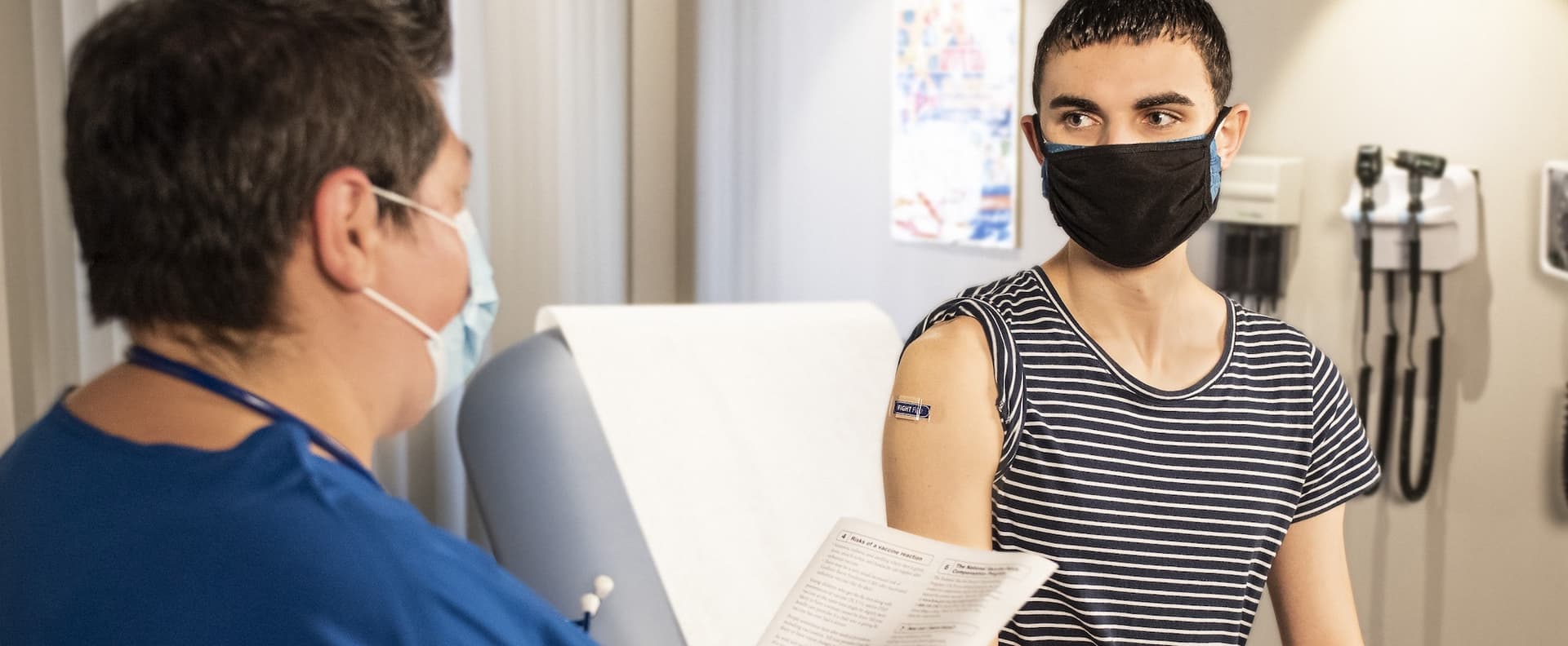 a double-masked patient listens to a nurse reading off of a vaccine information sheet