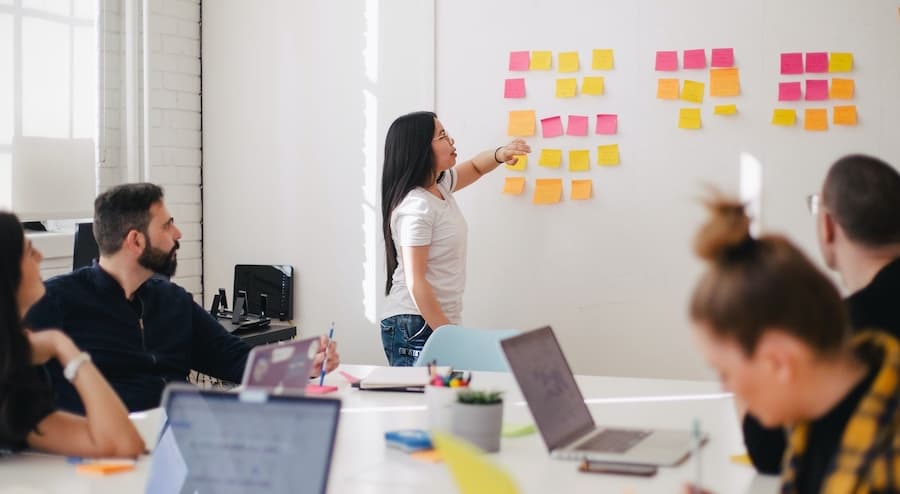 a woman in a meeting looking at a series of post it notes on a wall behind her with coworkers looking on with their laptops open on the table