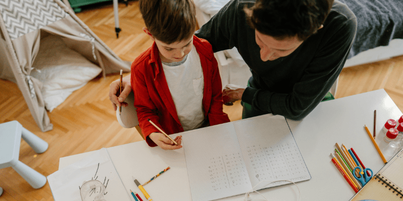 overhead shot of a parent looking over the shoulder of a child doing some math at a desk in their home