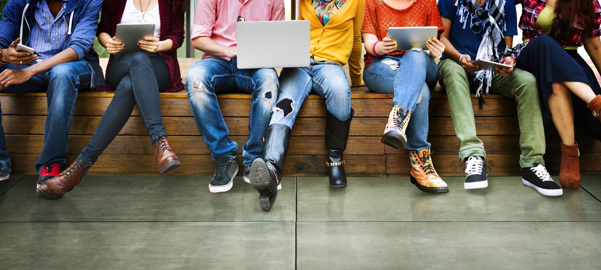 Group of people sitting next to each other holding different tech devices, from phones and ipads to computers.