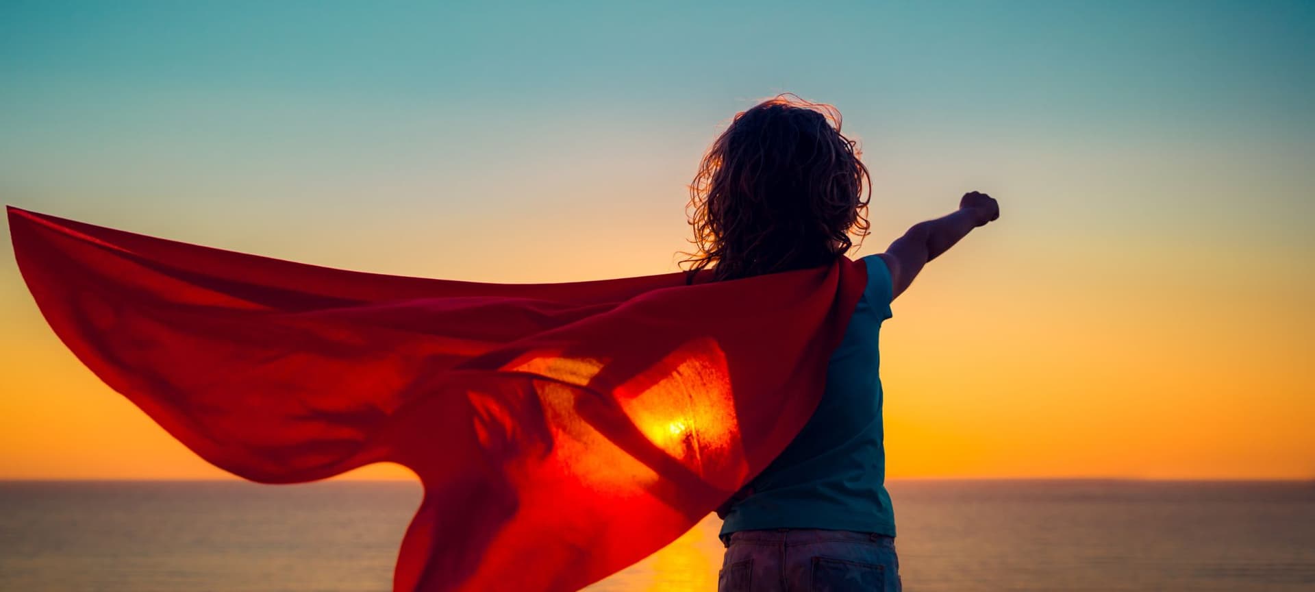 A child wearing a superhero cape posing like superman with arm extended with closed fist on a sunset lit beach