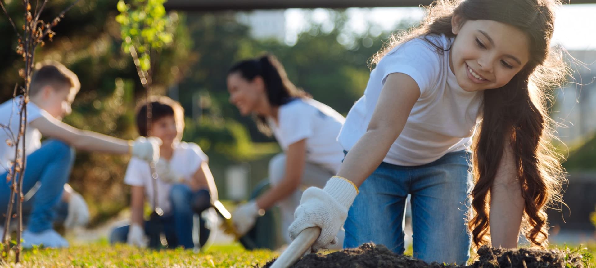 Family planting a tree in background, while young child tills the soil in preparation to plant another tree.