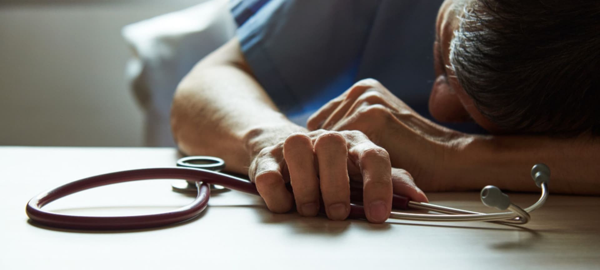 A medical doctor resting head on desk grasping his stethescope