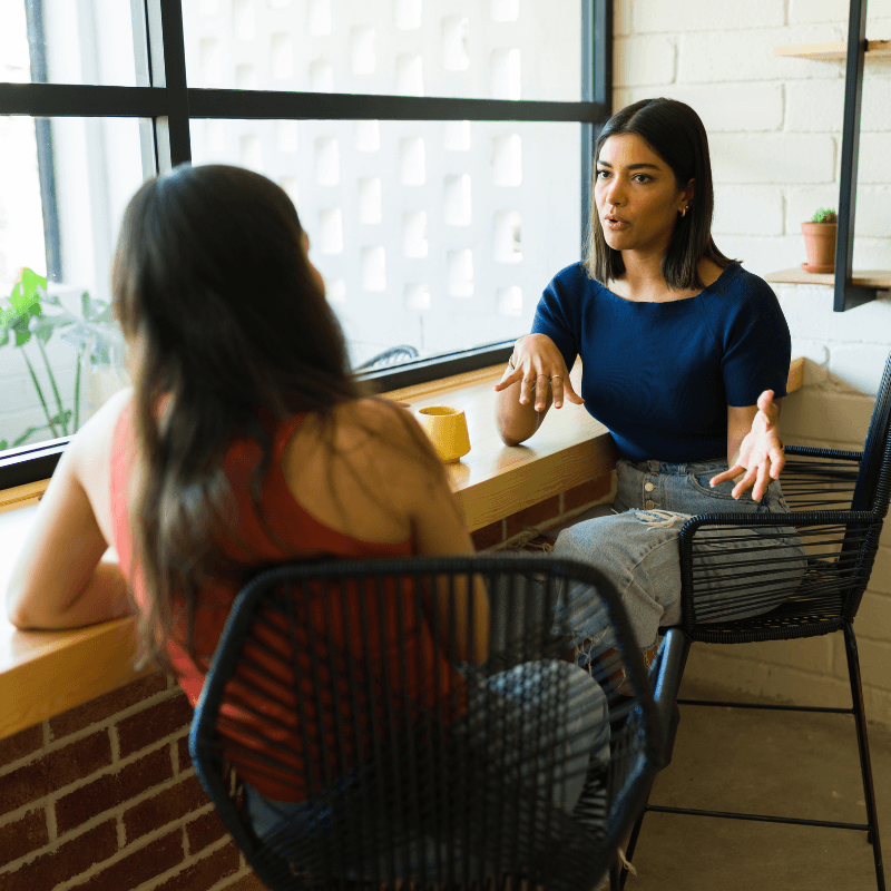Two women sitting in chairs in a coffee shop, having a conversation.