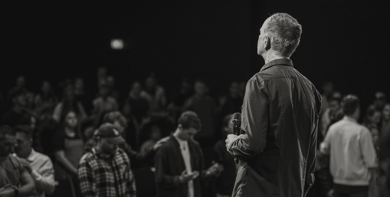 A pastor stands on a stage, holding a microphone and facing his congregation. The congregation bow their heads in prayer.