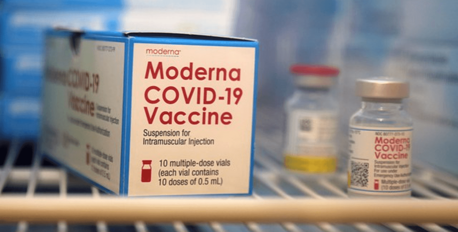 A box that reads "Moderna COVID-19 Vaccine" sits on a shelf in a refrigeration unit. Two vials of the vaccine sit next to it.