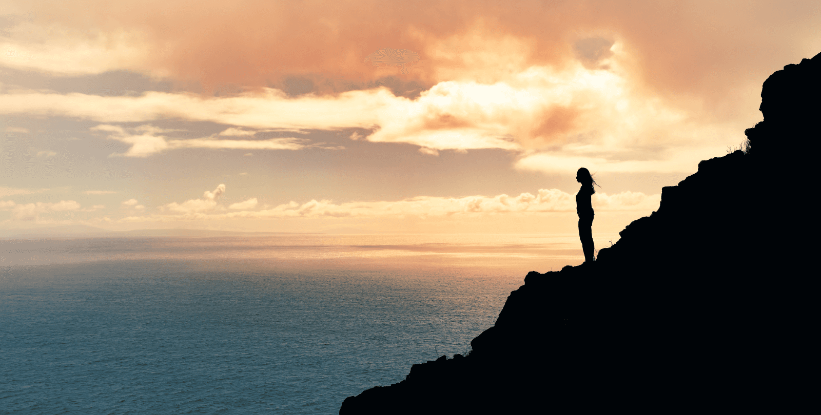 A woman, outlined in shadow, stands on the ridge of a mountain overlooking the ocean.