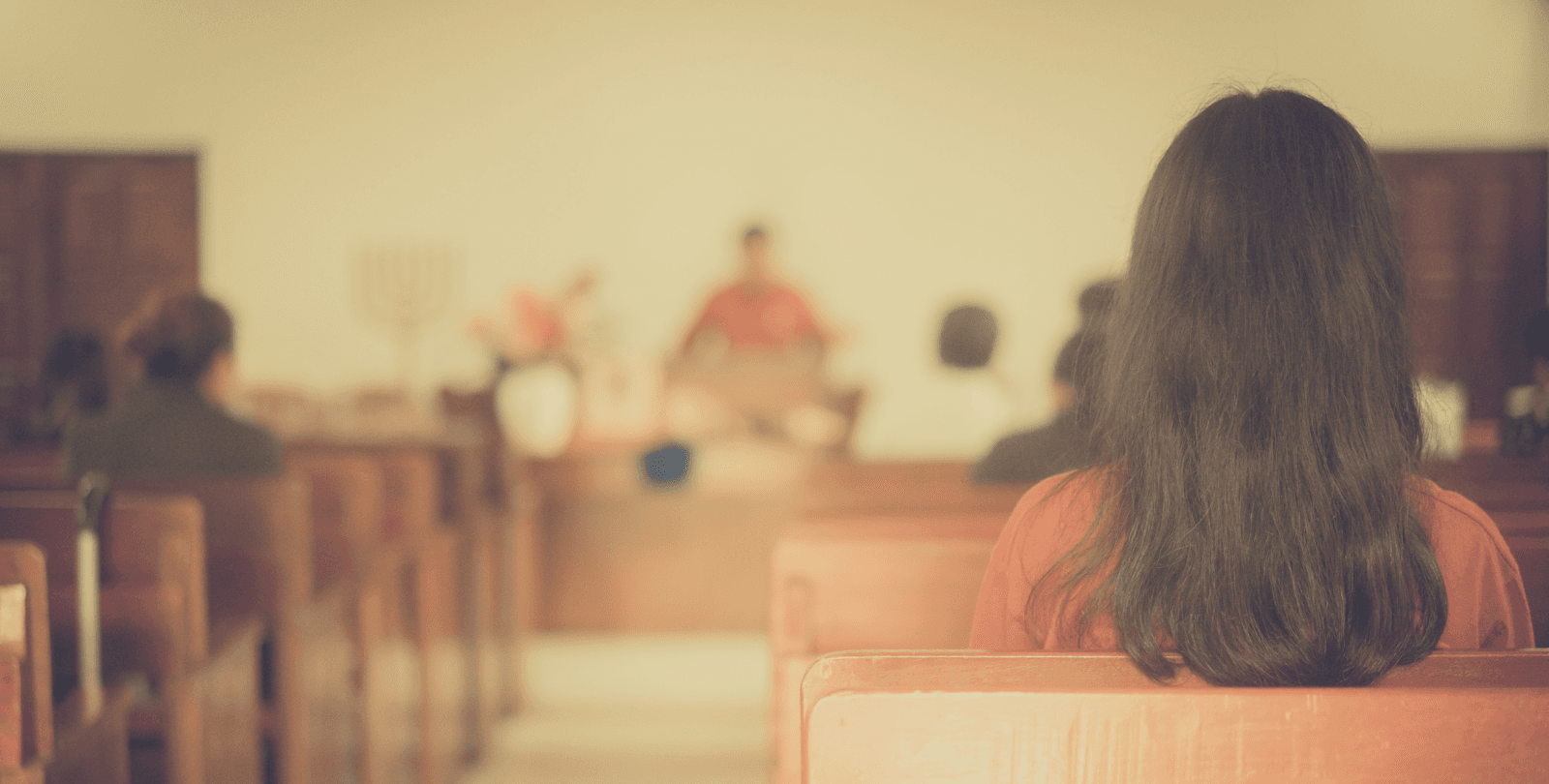 A woman sits in a pew and faces forward. A pastor stands at a pulpit and addresses the congregation.