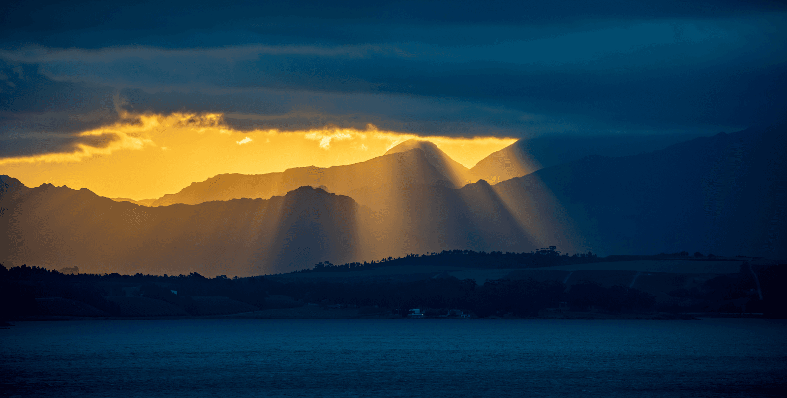 The Sun breaks through a patch of open sky surrounded by dark clouds. It shines down on mountain and water.