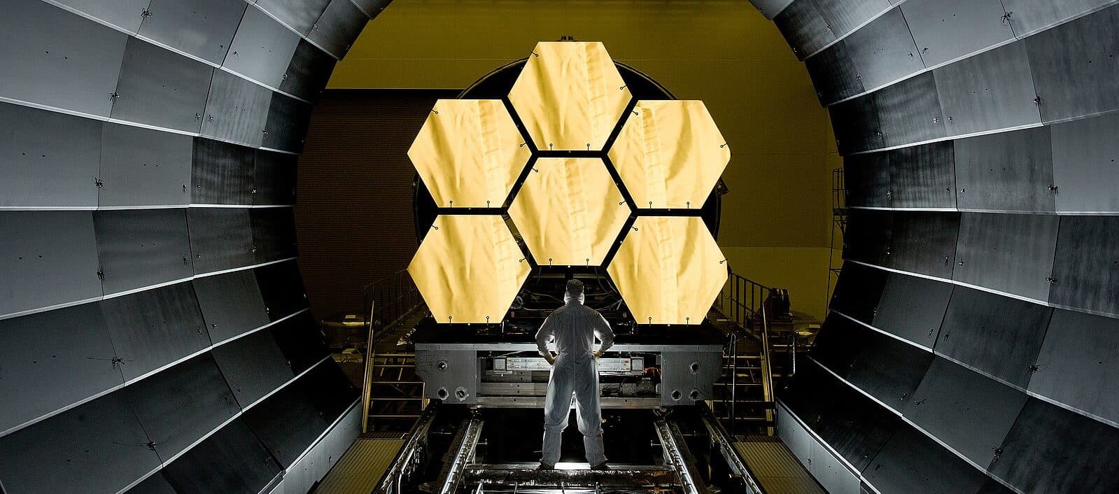 An engineer in a hazmat suit, with back to camera, stands in front of the James Webb Space Telescope's primary mirror system. The system is made up of six large, hexagonal mirrors. The mirror system faces into a large tunnel.