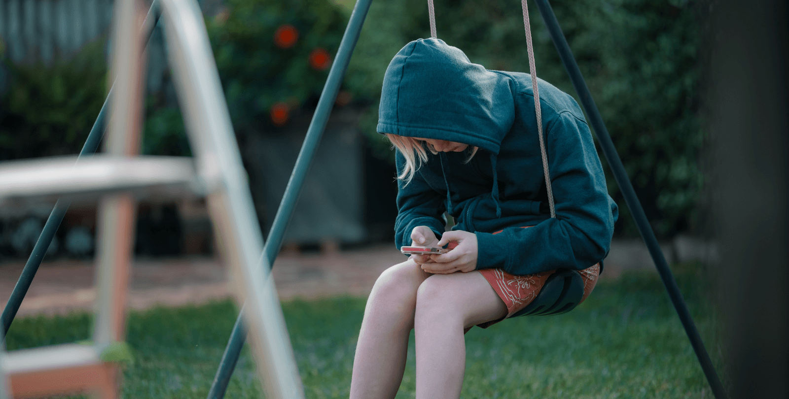 A teenager looks down at her phone as she sits on a playground swing.