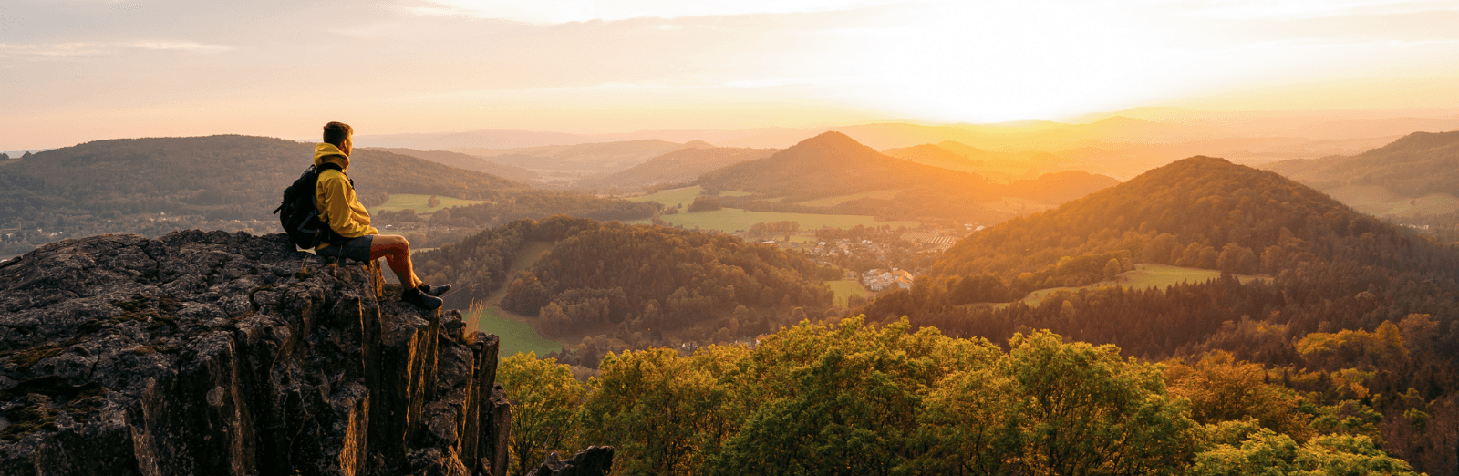 A man sits on the crest of a mountain and looks down at rolling valleys.