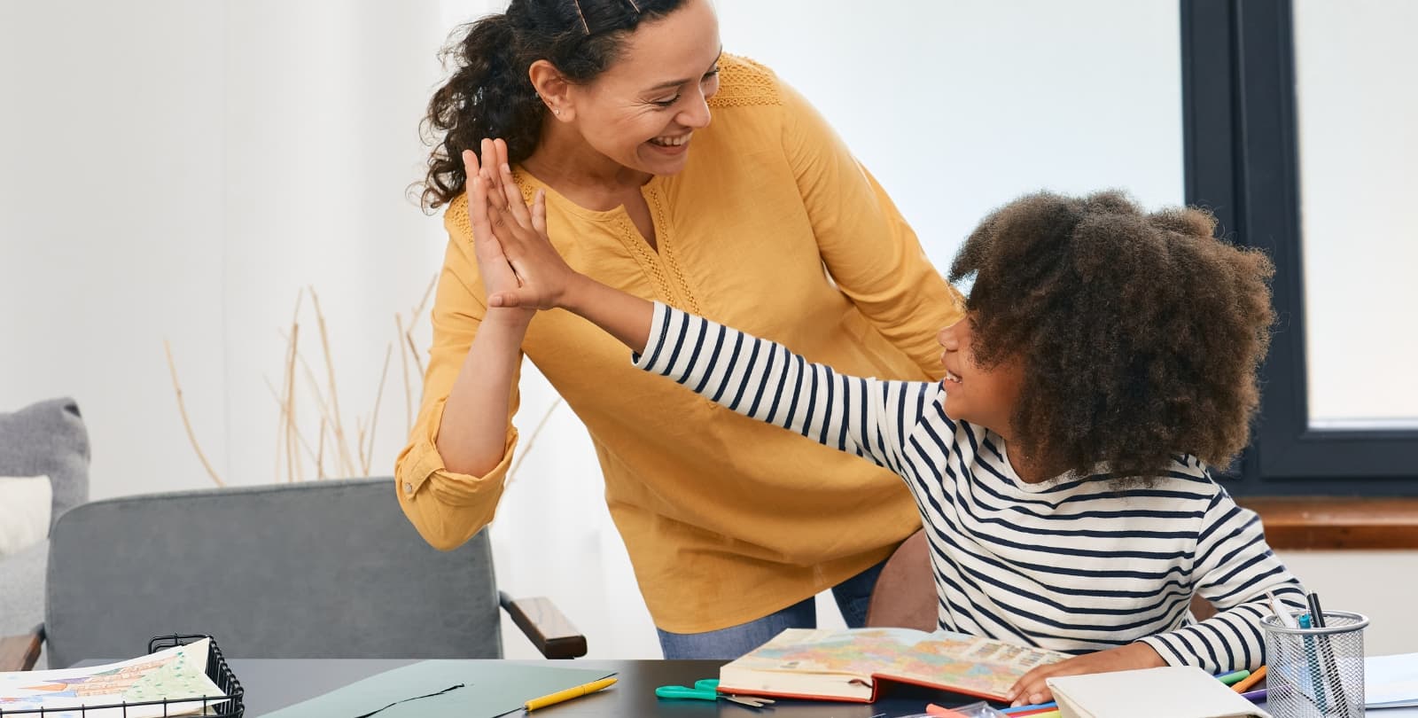 A child sits at a table covered in notebooks and pencils. She looks up at a woman and high fives her; both are smiling.