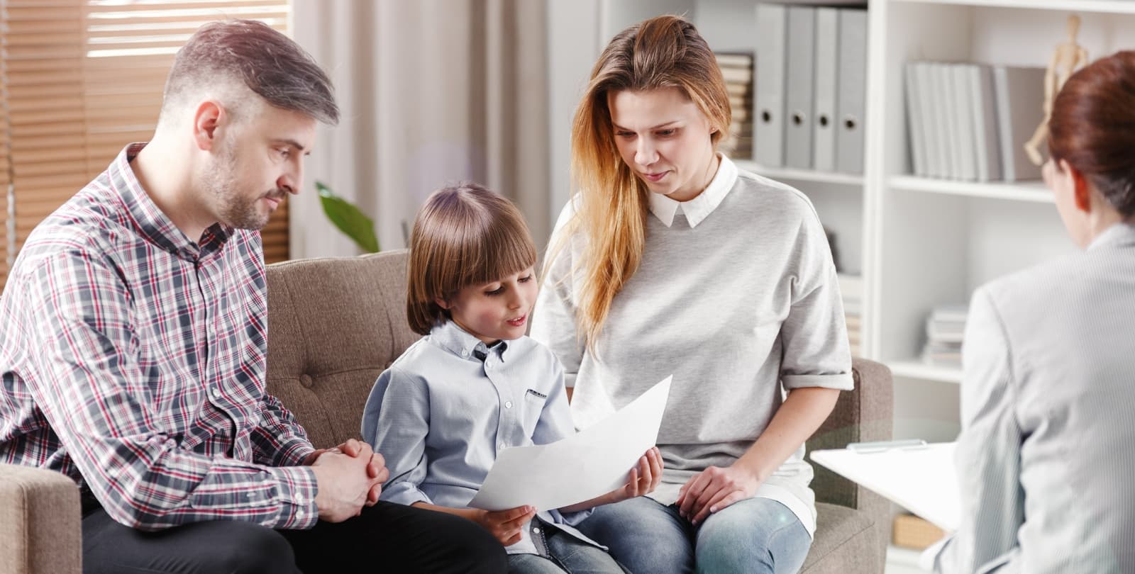 A child sits between his parents in the office of a mental health professional. The child and parents look down at a piece of paper. A therapist sits across from them.