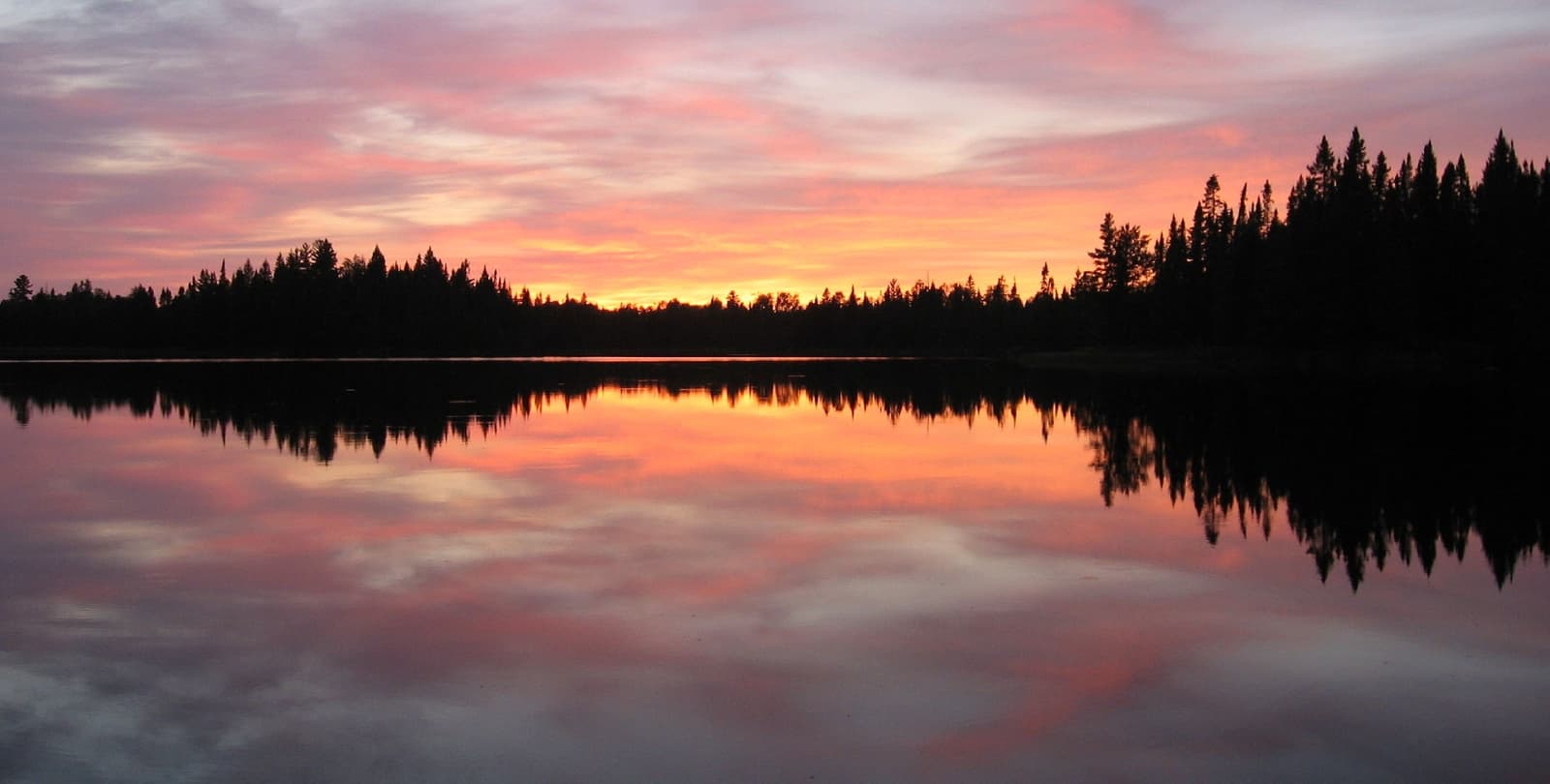 Sunset over a lake. Sunlight reflects off the still water. On the opposite shore, trees stand in shadow.