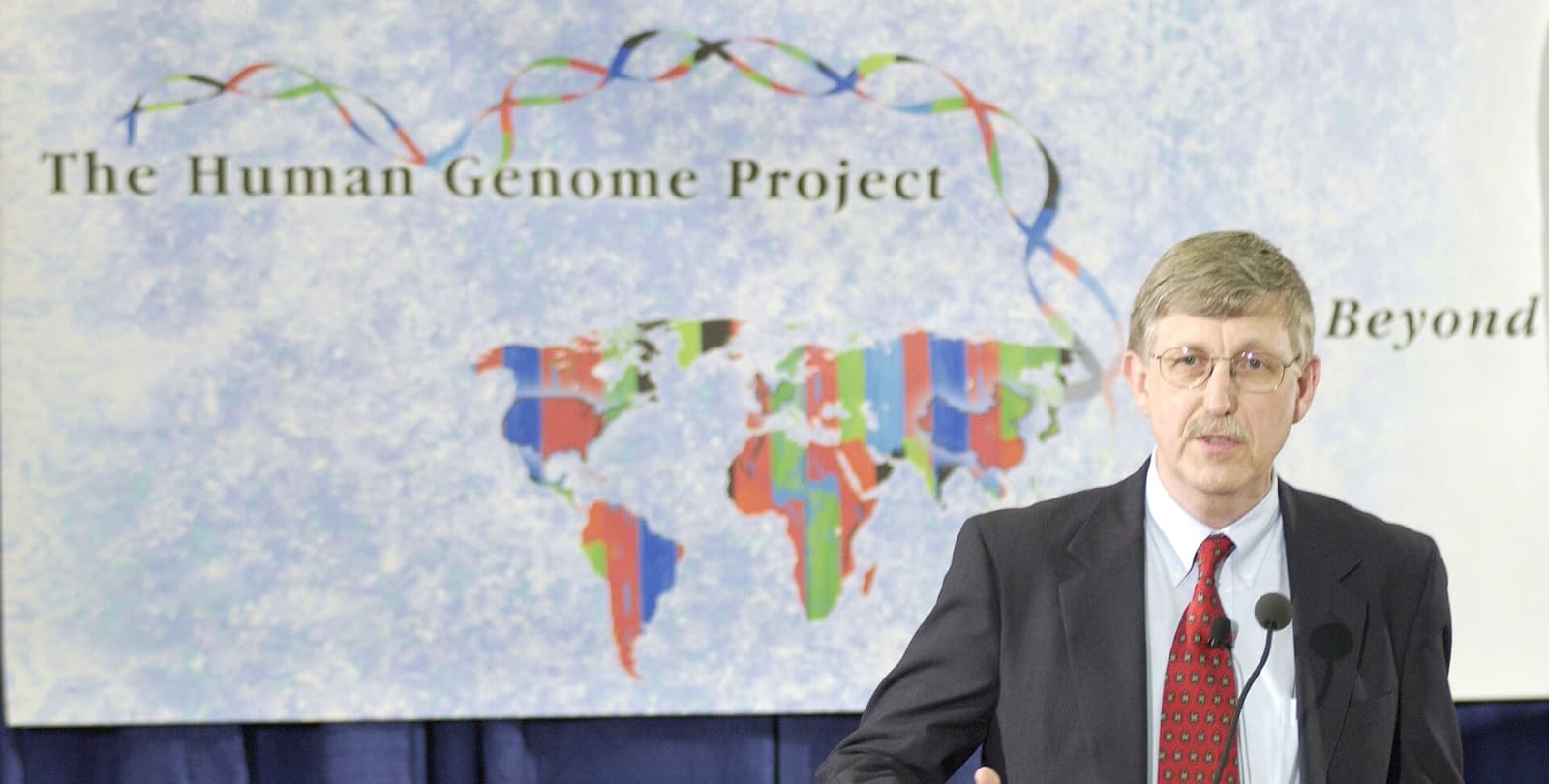 Francis Collins speaking at a podium in front of a sign that reads "The Human Genome Project."