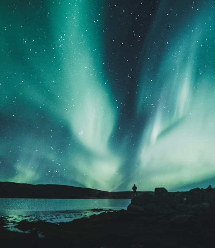 person standing near water looking at stars and northern lights