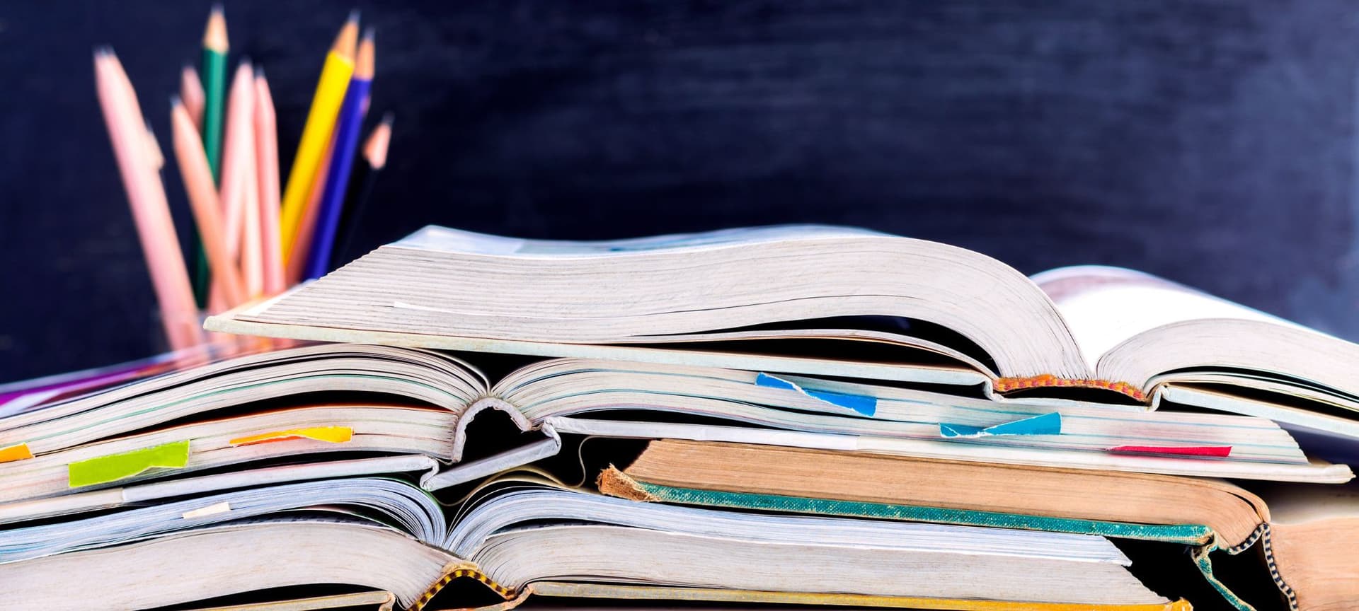 Open textbooks stacked on a desk in a classroom near blackboard and color pencils