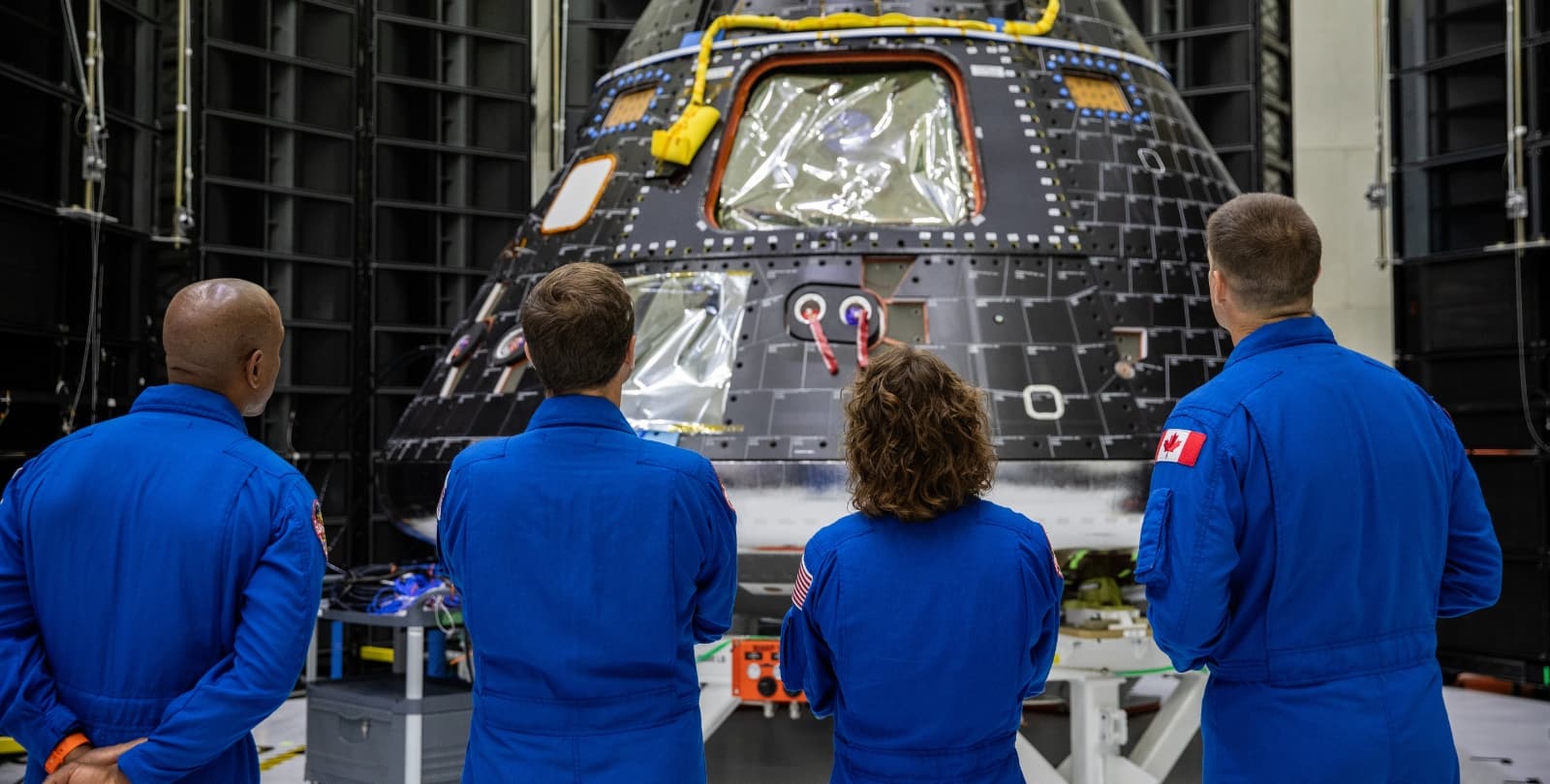 Four astronauts in jumpsuits stand and look at the Orion crew module. Their backs are to the camera.