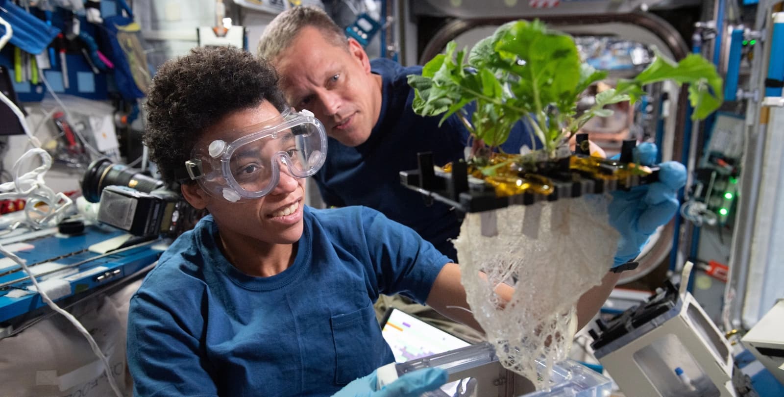 A NASA astronaut aboard the ISS holds a plant growth. Another astronaut watches over her shoulder.